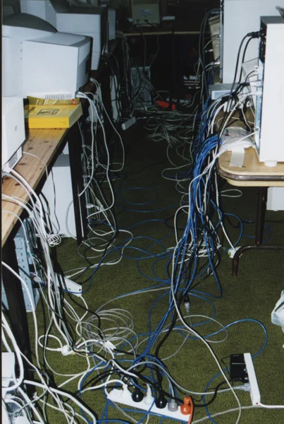 Under the tables at a 1998 LAN party: tangles of blue and white network cables and power boards across the carpet, beige PCs and CRT monitors on the desks above